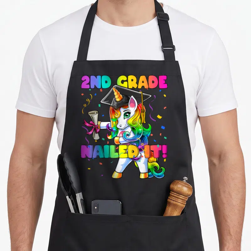 Flossing 2nd apron - Cartoon unicorn with rainbow mane holding diploma under 2ND GRADE NAILED IT! text — shown on a black cotton canvas apron against a white background