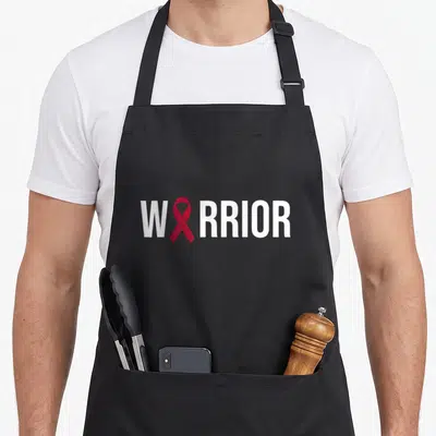 Multiple myeloma ribbon apron - WARRIOR text with red ribbon replacing the letter — shown on a black cotton canvas apron against a white background