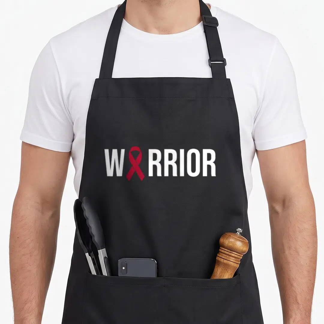 Multiple myeloma ribbon apron - WARRIOR text with red ribbon replacing the letter — shown on a black cotton canvas apron against a white background