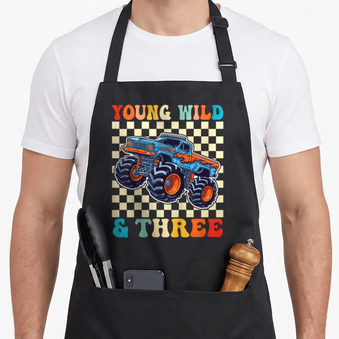 Young truck apron - Blue monster truck over cream checkerboard with colorful YOUNG WILD & THREE text — shown on a black cotton canvas apron against a white background