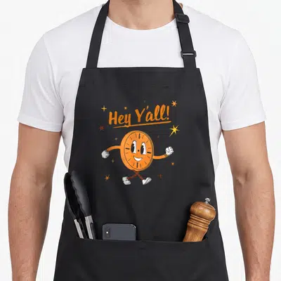 Minute little miss apron - Walking orange clock character with Hey Y'all text and scattered stars in cartoon style — shown on a black cotton canvas apron against a white background