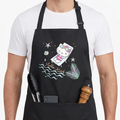 Funny summer aprons - Hello Kitty in pink bikini on striped beach chair with starfish shells and waves — shown on a black cotton canvas apron against a white background