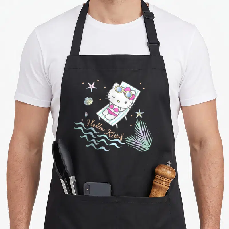 Funny summer aprons - Hello Kitty in pink bikini on striped beach chair with starfish shells and waves — shown on a black cotton canvas apron against a white background