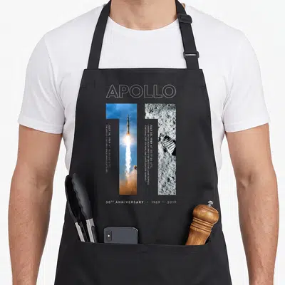 Landing apollo apron - Apollo 11 50th anniversary print with launch and boot print photo strips and white outline text — shown on a black cotton canvas apron against a white background