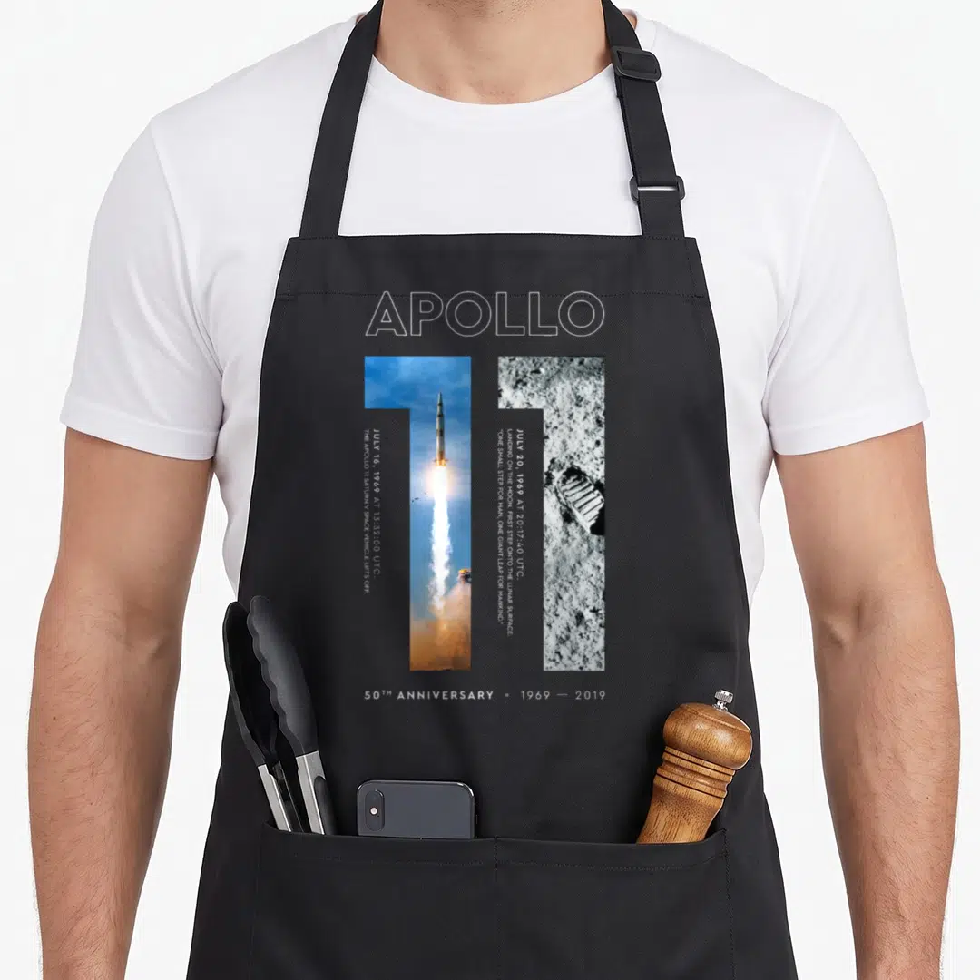 Landing apollo apron - Apollo 11 50th anniversary print with launch and boot print photo strips and white outline text — shown on a black cotton canvas apron against a white background