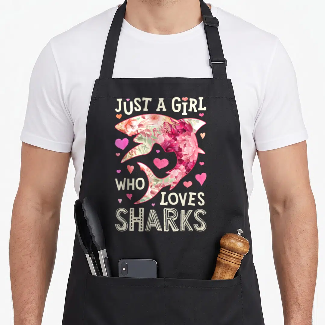 Shark floral apron - Floral filled shark silhouette with scattered hearts and text Just A Girl Who Loves Sharks — shown on a black cotton canvas apron against a white background