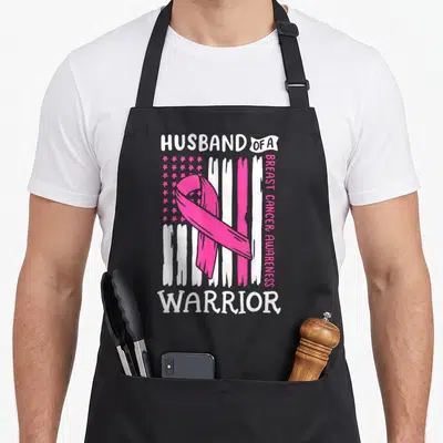 Warrior support squad breast cancer awareness aprons - Husband of a Warrior text with pink ribbon and distressed flag — shown on a black cotton canvas apron against a white background