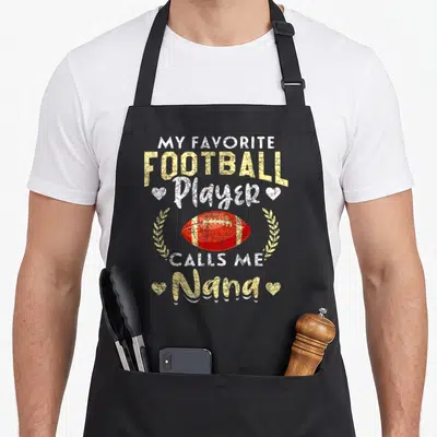 Retired grandma aprons - Distressed text My Favorite Football Player Calls Me Nana with red football and yellow wreath — shown on a black cotton canvas apron against a white background