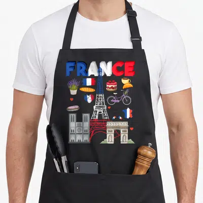 France chef outfit with apron - Cartoon Eiffel Tower surrounded by FRANCE text, lavender, croissants, and Paris — shown on a black cotton canvas apron against a white background