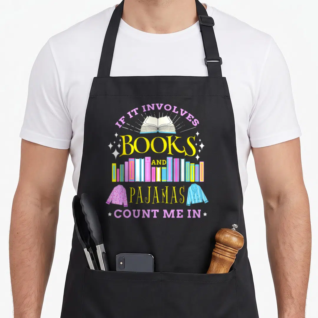 Involve hiking lover apron - Open book, colorful book row, pink and blue pajama jackets, white stars, printed text — shown on a black cotton canvas apron against a white background