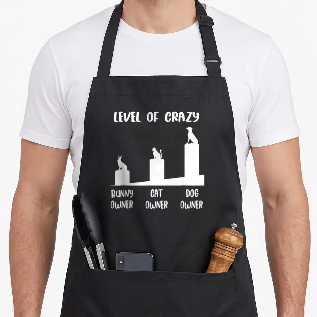 Owner cooking with dog apron - White bar chart showing bunny, cat, and dog owner silhouettes with text Level of Crazy — shown on a black cotton canvas apron against a white background