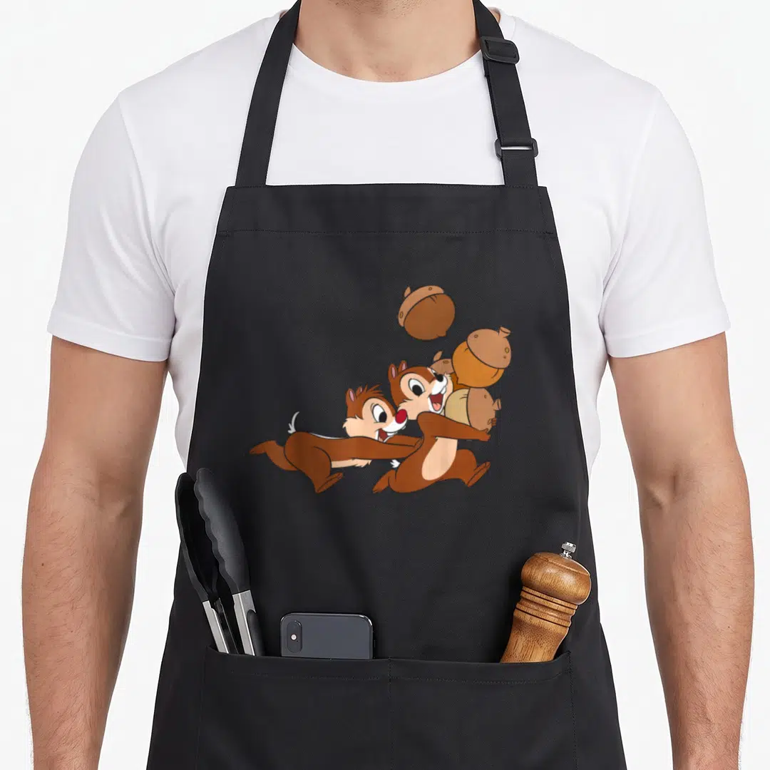 Chip acorns and aprons - Two cartoon chipmunks in brown and tan holding acorns with one floating above — shown on a black cotton canvas apron against a white background