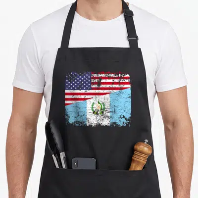 Guatemalan root american flag apron - Two flags merged diagonally in distressed red white blue and light blue — shown on a black cotton canvas apron against a white background