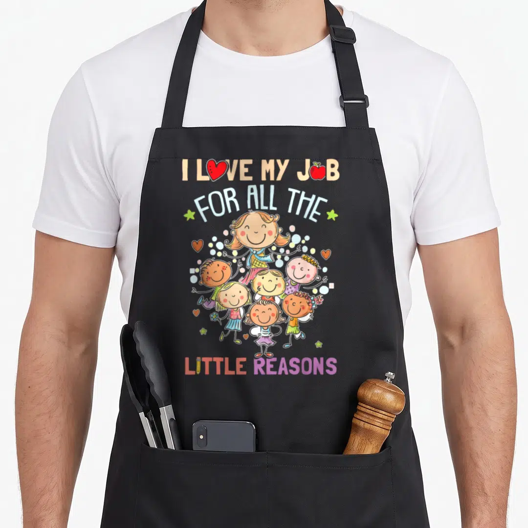 Teaching teacher apron - Text I Love My Job surrounded by cartoon children, hearts, stars, and dots in pink red yellow — shown on a black cotton canvas apron against a white background