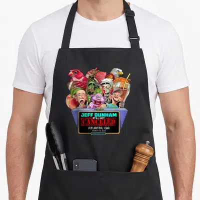 Jeff trailer guy apron - Cartoon Jeff Dunham puppets and sports gear on blue podium with Still Not Canceled text — shown on a black cotton canvas apron against a white background