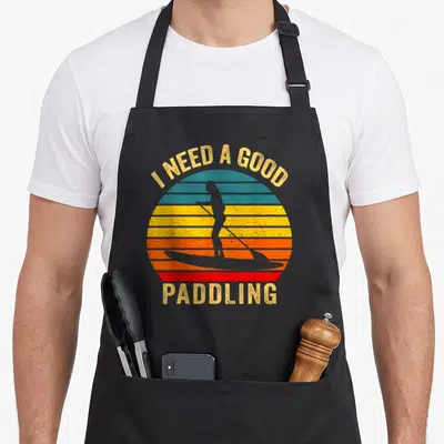 Paddling paddle apron - Retro striped sun with paddleboarder silhouette and text I NEED A GOOD PADDLING — shown on a black cotton canvas apron against a white background