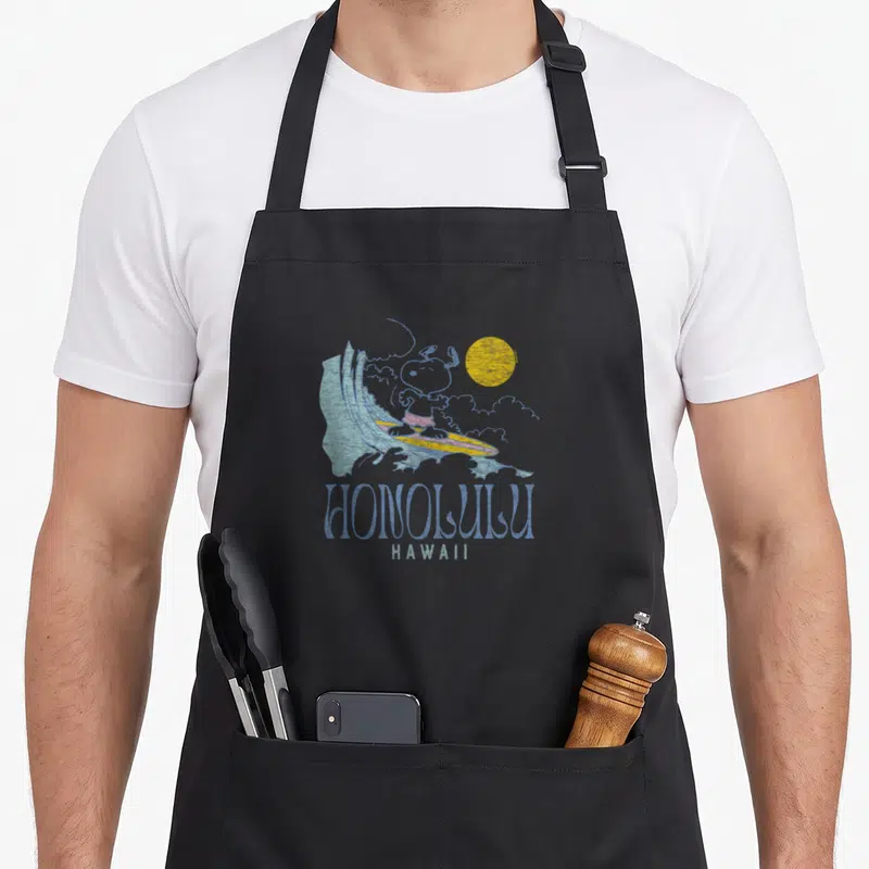 Surfing hawaiian apron - Distressed Snoopy surfing wave with blue HONOLULU HAWAII text below — shown on a black cotton canvas apron against a white background