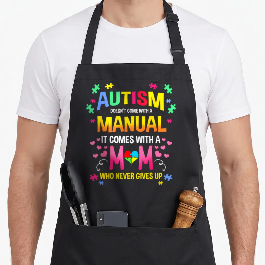 Manual autism apron - Colorful stacked text reading autism manual mom with puzzle pieces and heart graphics — shown on a black cotton canvas apron against a white background