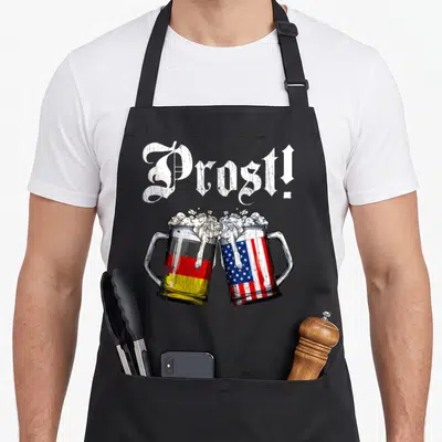 Prost beer apron - Two clinking beer mugs wrapped in German and American flags under distressed Prost text — shown on a black cotton canvas apron against a white background