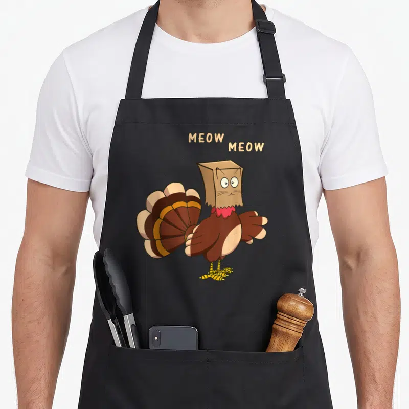 Cat turkey apron - Cartoon turkey in paper bag cat mask with two MEOW text bubbles above head — shown on a black cotton canvas apron against a white background