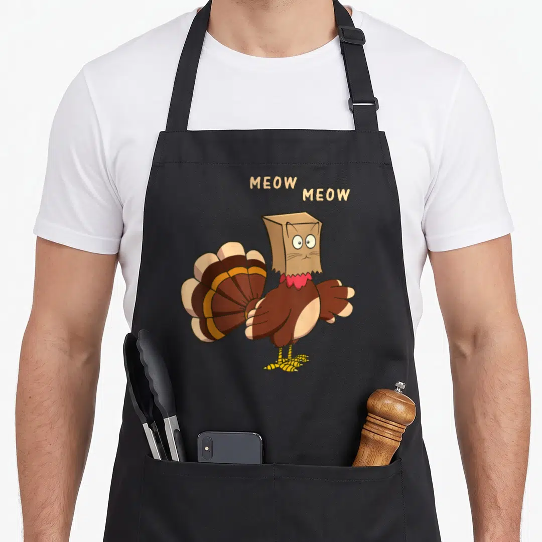Cat turkey apron - Cartoon turkey in paper bag cat mask with two MEOW text bubbles above head — shown on a black cotton canvas apron against a white background