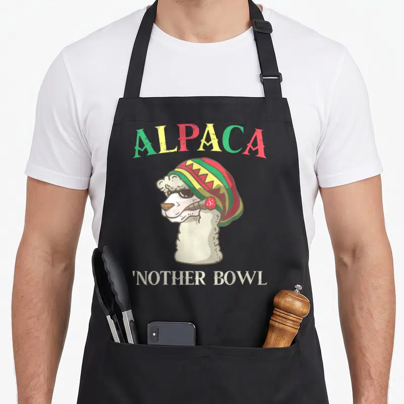 Grass alpaca apron - Cartoon alpaca in rasta beanie smoking with arched text ALPACA and bottom text Nother Bowl — shown on a black cotton canvas apron against a white background