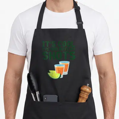 Both funny tequila apron - Green text IT'S COOL I'VE HAD BOTH MY SHOTS above two orange shot glasses and two lime slices — shown on a black cotton canvas apron against a white background