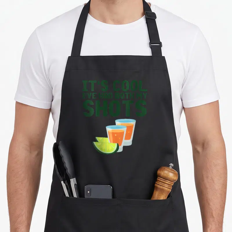 Both funny tequila apron - Green text IT'S COOL I'VE HAD BOTH MY SHOTS above two orange shot glasses and two lime slices — shown on a black cotton canvas apron against a white background