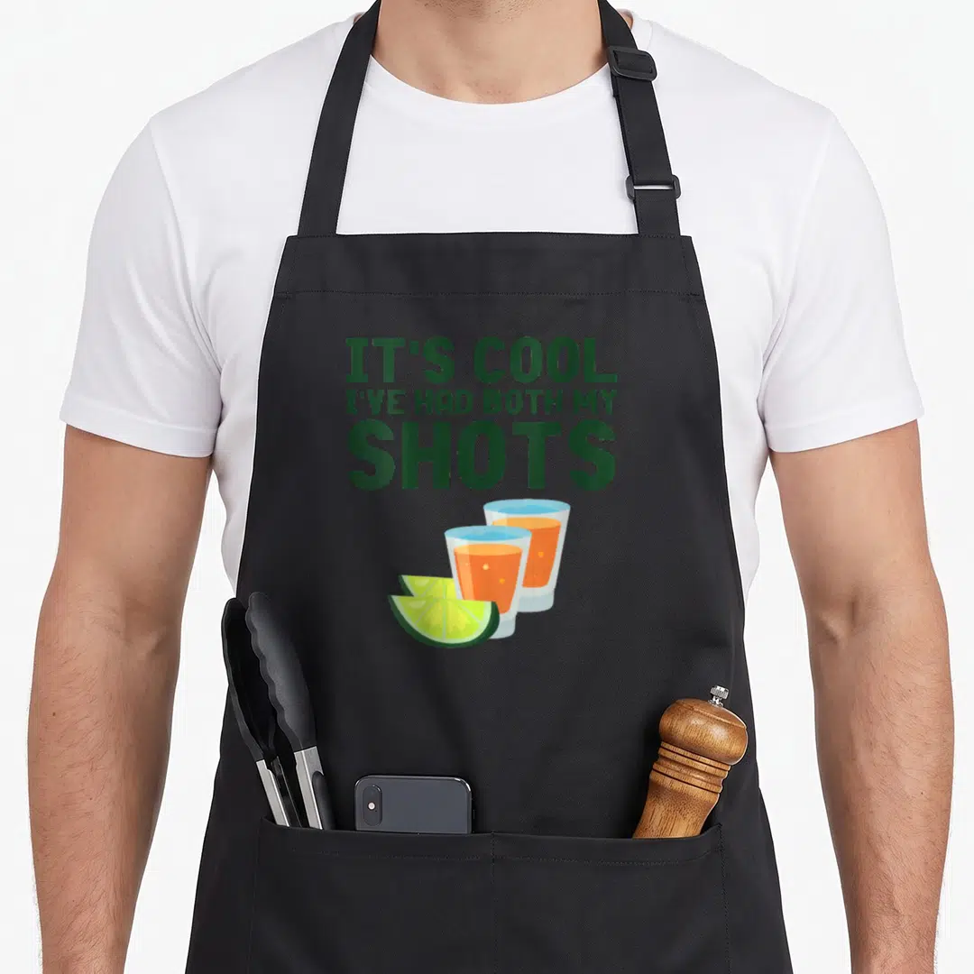 Both funny tequila apron - Green text IT'S COOL I'VE HAD BOTH MY SHOTS above two orange shot glasses and two lime slices — shown on a black cotton canvas apron against a white background