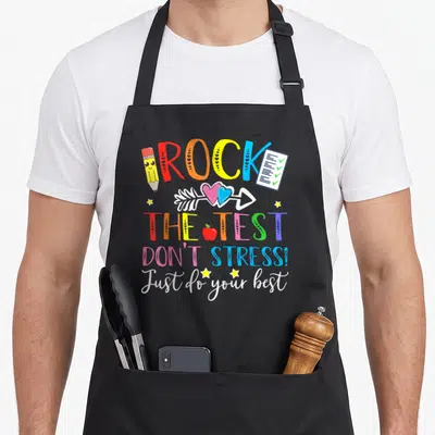Teacher test kitchen apron - Colorful cartoon text ROCK THE TEST with pencil checklist apple and hearts — shown on a black cotton canvas apron against a white background