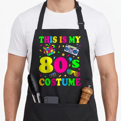 Das 80s apron - Colorful 80s text with Rubik's cube, boombox, and shutter shades — shown on a black cotton canvas apron against a white background
