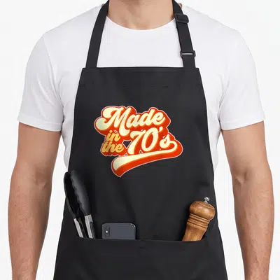 Funny 70s apron - Retro orange and cream typography reading Made in the 70s with a swoosh underline — shown on a black cotton canvas apron against a white background