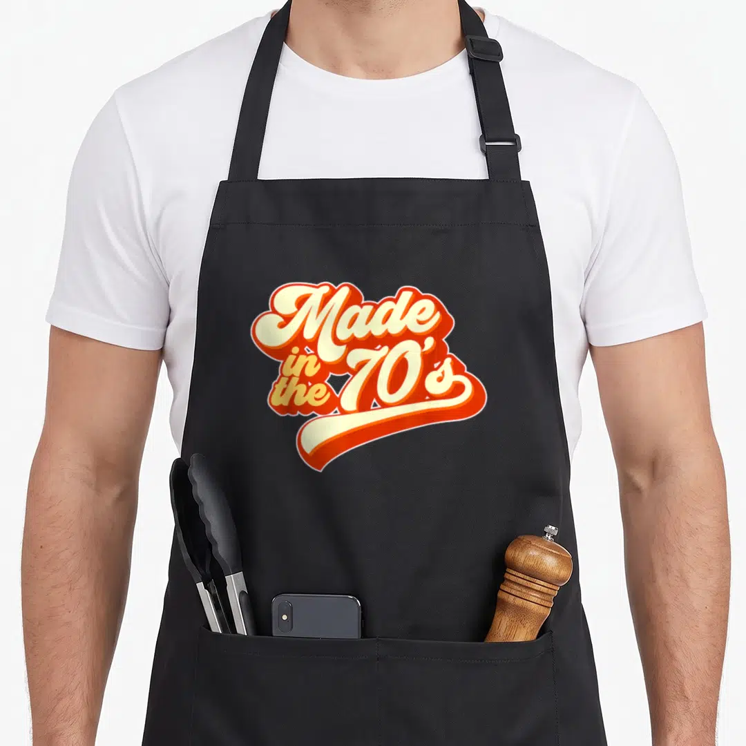 Funny 70s apron - Retro orange and cream typography reading Made in the 70s with a swoosh underline — shown on a black cotton canvas apron against a white background