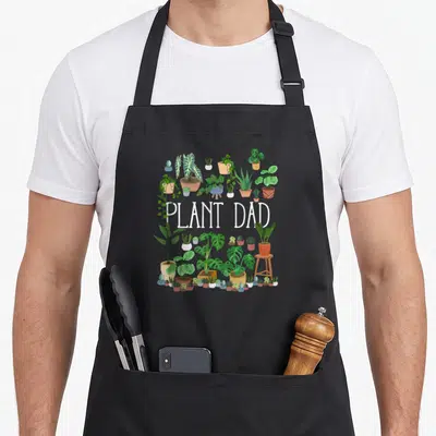 Potted plant apron - PLANT DAD text surrounded by green houseplants in white and brown pots with a small stool — shown on a black cotton canvas apron against a white background