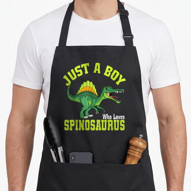 Funny spinosaurus apron - Cartoon green spinosaurus with arched text Just A Boy Who Loves Spinosaurus — shown on a black cotton canvas apron against a white background