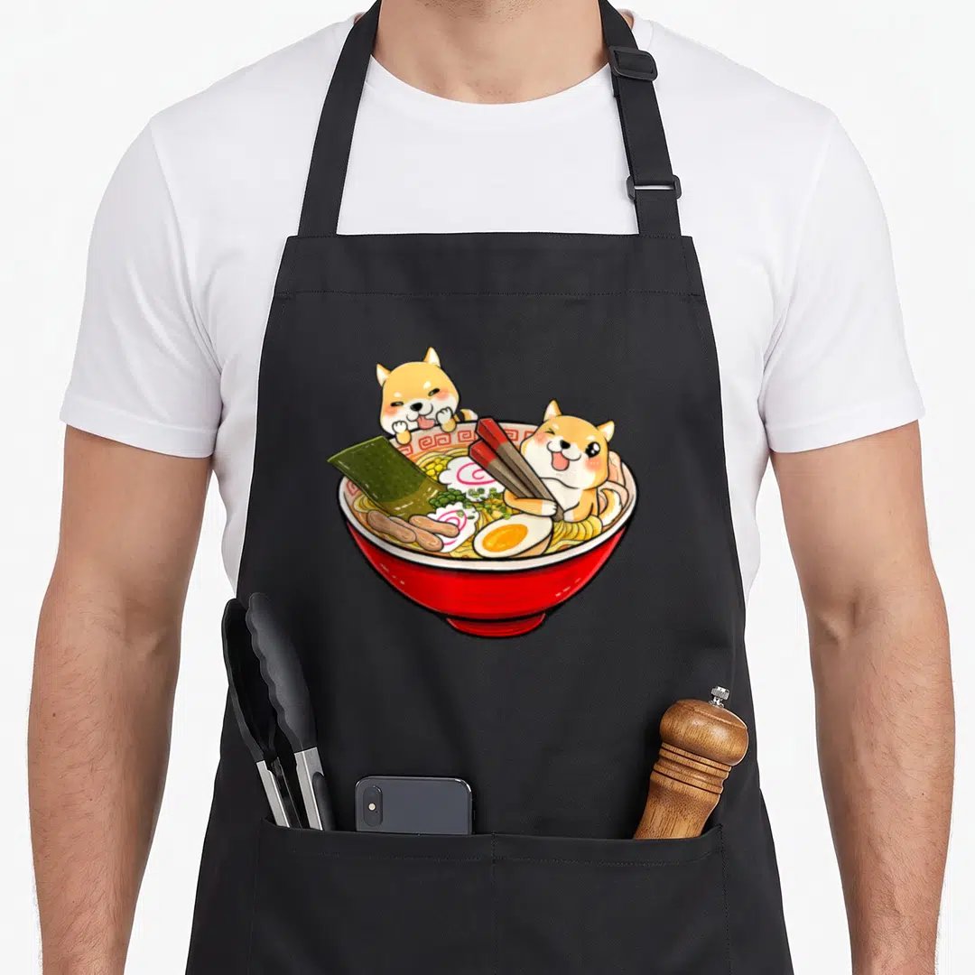 Inu japanese dog apron - Two shiba inu dogs sitting in a red ramen bowl with noodles, egg, and chopsticks — shown on a black cotton canvas apron against a white background