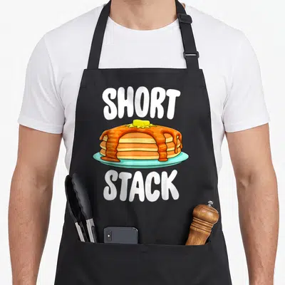 Foodie pancake apron - Cartoon pancakes with butter on blue plate surrounded by white text reading short stack — shown on a black cotton canvas apron against a white background