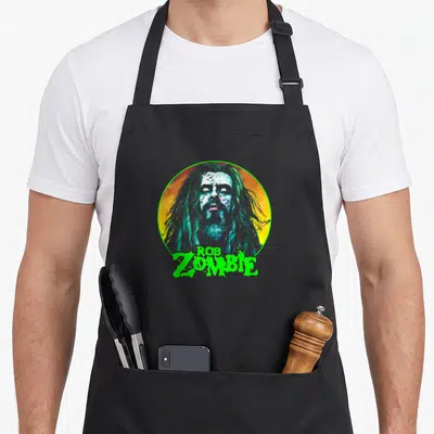 Rob face apron - Cartoon zombie face in yellow orange circle with green Rob Zombie text below — shown on a black cotton canvas apron against a white background