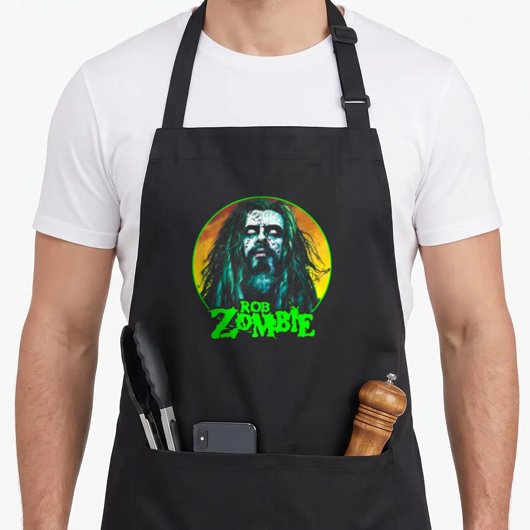 Rob face apron - Cartoon zombie face in yellow orange circle with green Rob Zombie text below — shown on a black cotton canvas apron against a white background