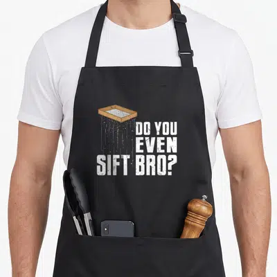 Even archaeology hobby apron - Brown sifting tray with falling white particles next to distressed white stacked text DO — shown on a black cotton canvas apron against a white background