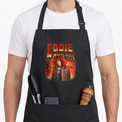 Eddie stranger apron - Eddie Munson portrait with fire lightning and Hellfire Club logo in red orange yellow black — shown on a black cotton canvas apron against a white background