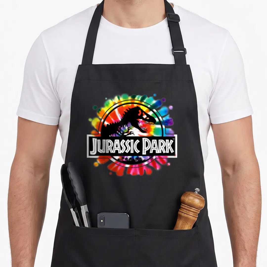 Classis tie apron - Tie-dye Jurassic Park logo with black T-Rex silhouette, white text banner, and palm tree — shown on a black cotton canvas apron against a white background