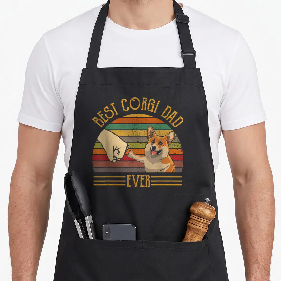 Sunset corgi apron - Retro corgi fist-bumping human hand inside striped sunset circle with orange and yellow text — shown on a black cotton canvas apron against a white background