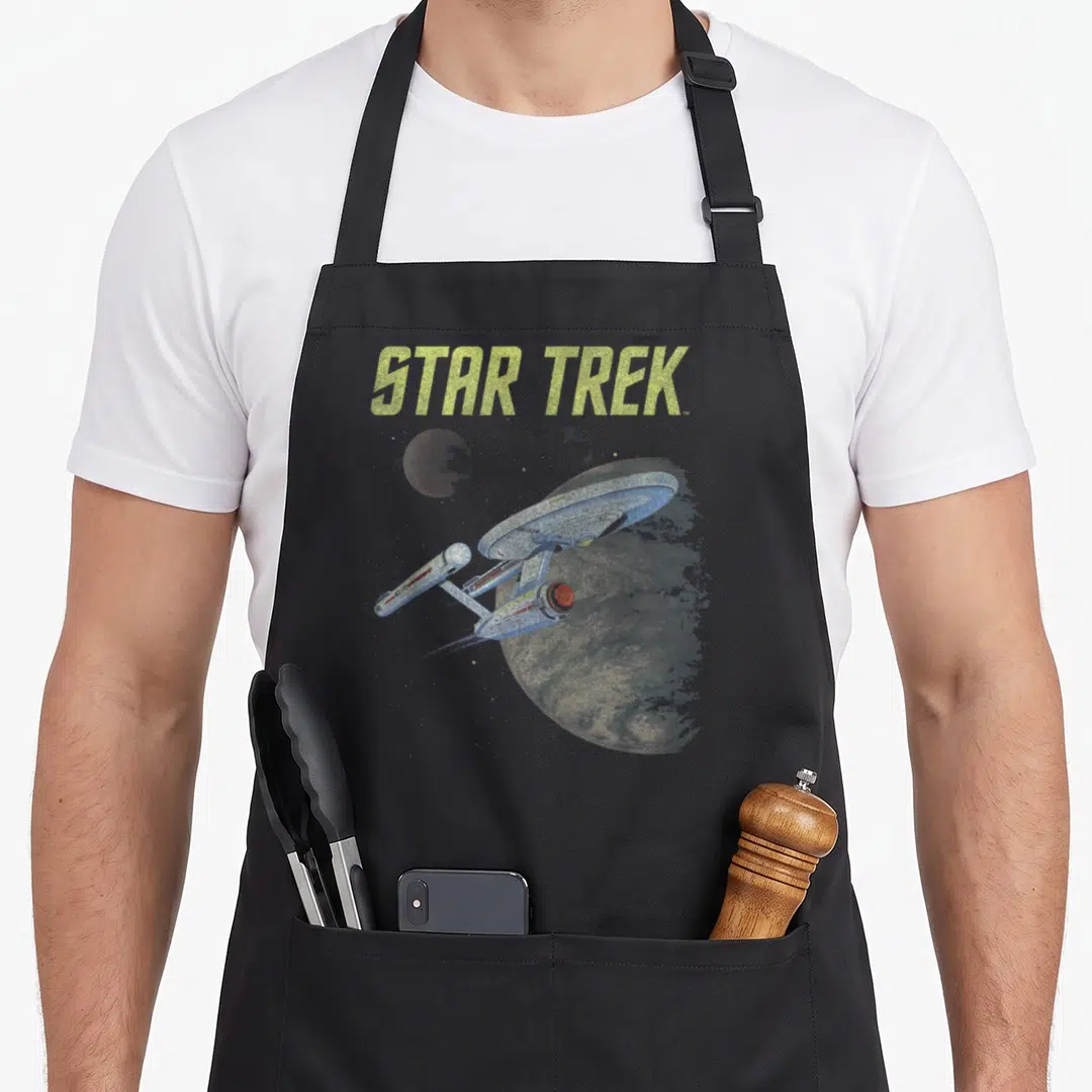 Enterprise star apron - STAR TREK text above spaceship flying past planets in distressed yellow and grey — shown on a black cotton canvas apron against a white background