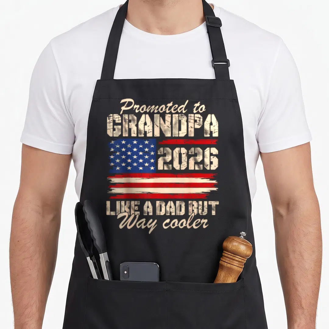 Est grandad aprons - Distressed Promoted to GRANDPA 2026 text with overlapping blue red white USA flag graphic — shown on a black cotton canvas apron against a white background