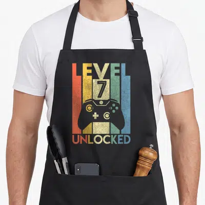 7th birthday apron - Retro game controller silhouette between striped LEVEL 7 UNLOCKED text in red yellow beige green — shown on a black cotton canvas apron against a white background