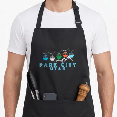 Park first snow apron - Five colorful gondola cars above white peaks with Park City Utah text below — shown on a black cotton canvas apron against a white background