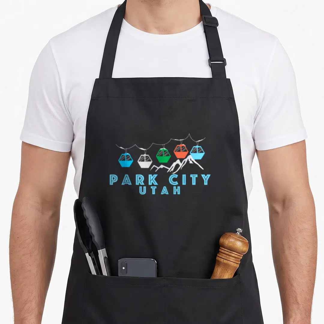 Park first snow apron - Five colorful gondola cars above white peaks with Park City Utah text below — shown on a black cotton canvas apron against a white background