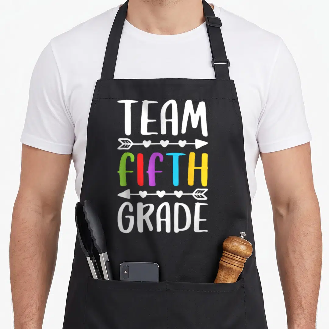 Fifth grade teacher apron - Stacked TEAM FIFTH GRADE text with colorful letters and heart arrows on black fabric — shown on a black cotton canvas apron against a white background