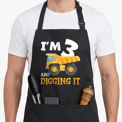 Three birthday apron - Cartoon yellow dump truck with white and orange text reading I'M 3 AND DIGGING IT on grey — shown on a black cotton canvas apron against a white background